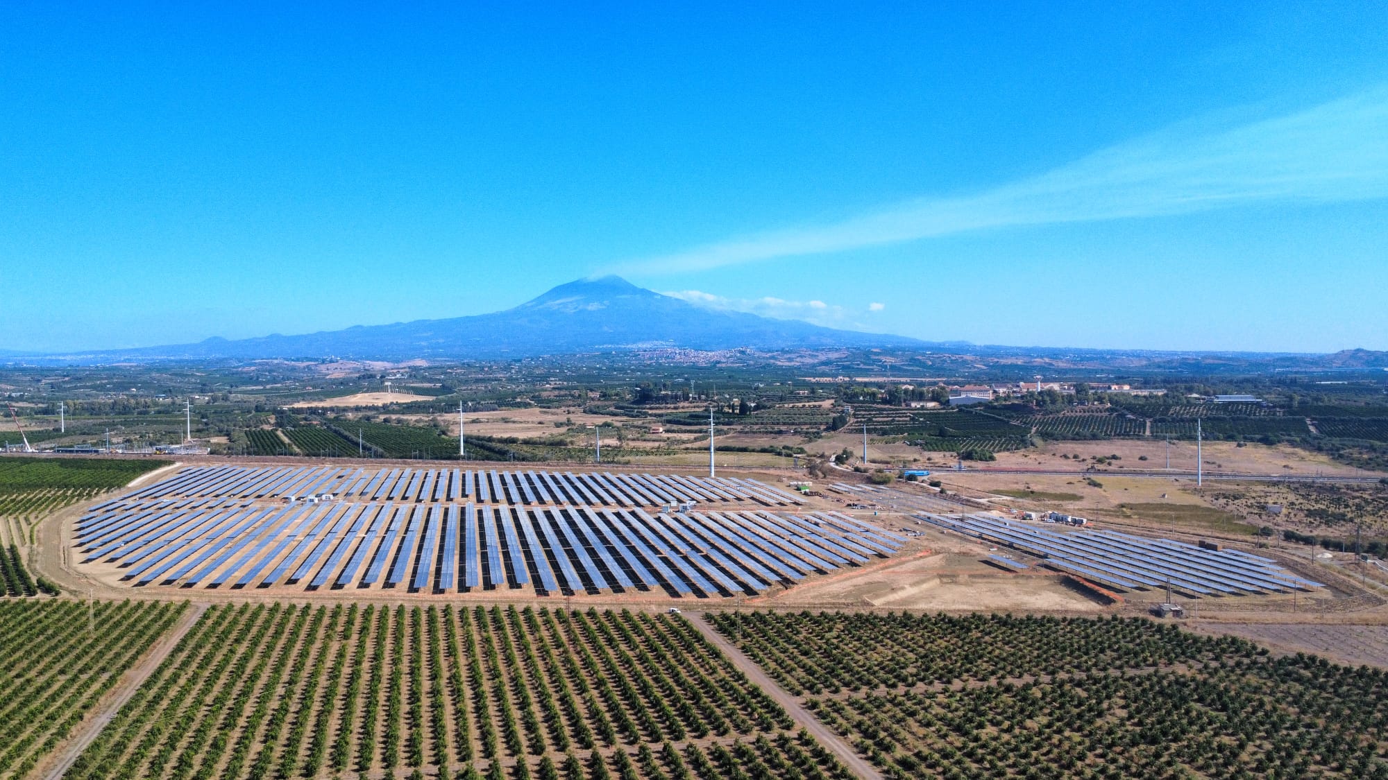 Aerial view of a large photovoltaic plant located in a rural area, surrounded by cultivated fields. In the background, Mount Etna rises under a clear blue sky.