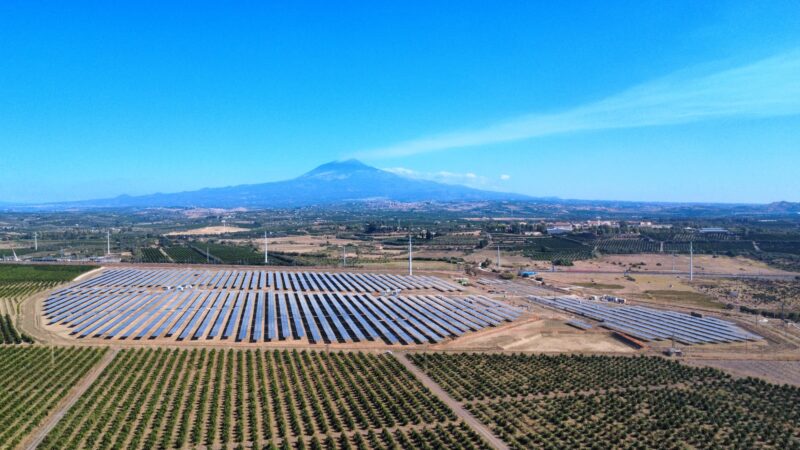 Veduta aerea di un grande impianto fotovoltaico situato in una zona rurale, circondato da campi coltivati. Sullo sfondo si erge l'Etna sotto un cielo limpido e azzurro.
